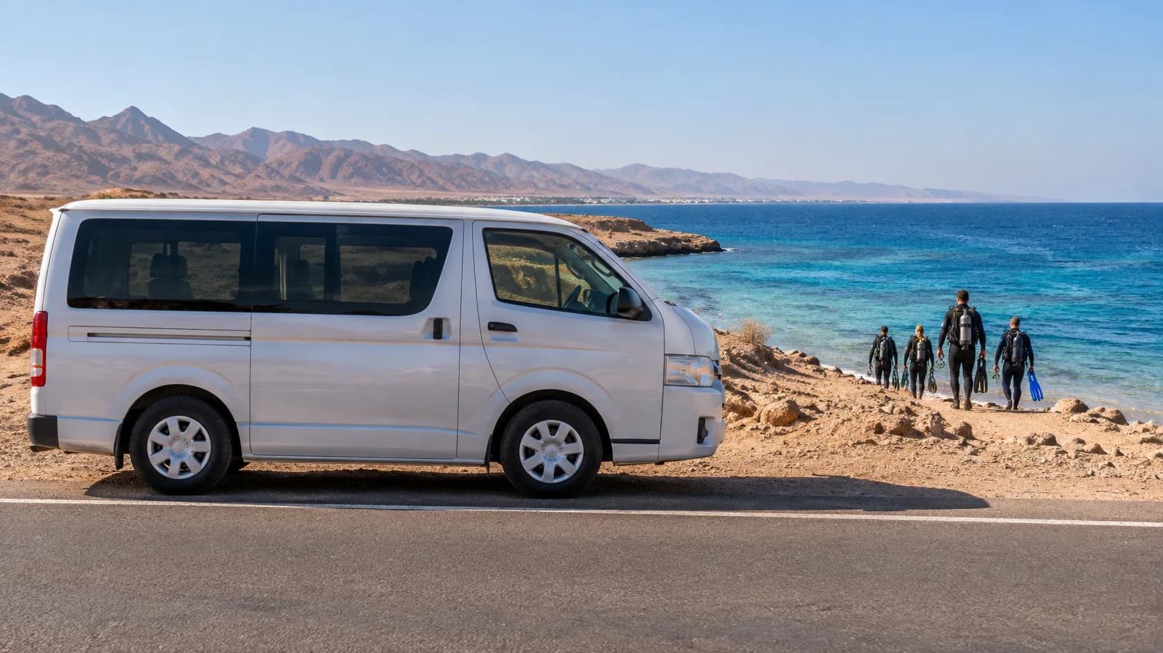 White passenger van parked beside a coastal road, with five divers carrying gear walking toward the blue sea and mountains